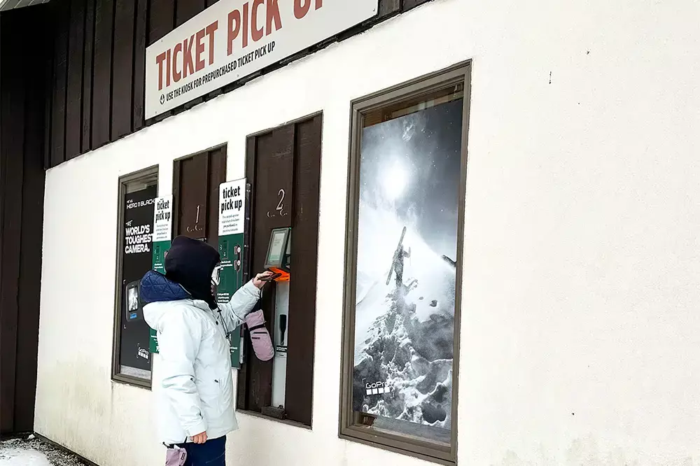 Person using a ticket pickup kiosk at a snowy location, with mountain photography nearby.