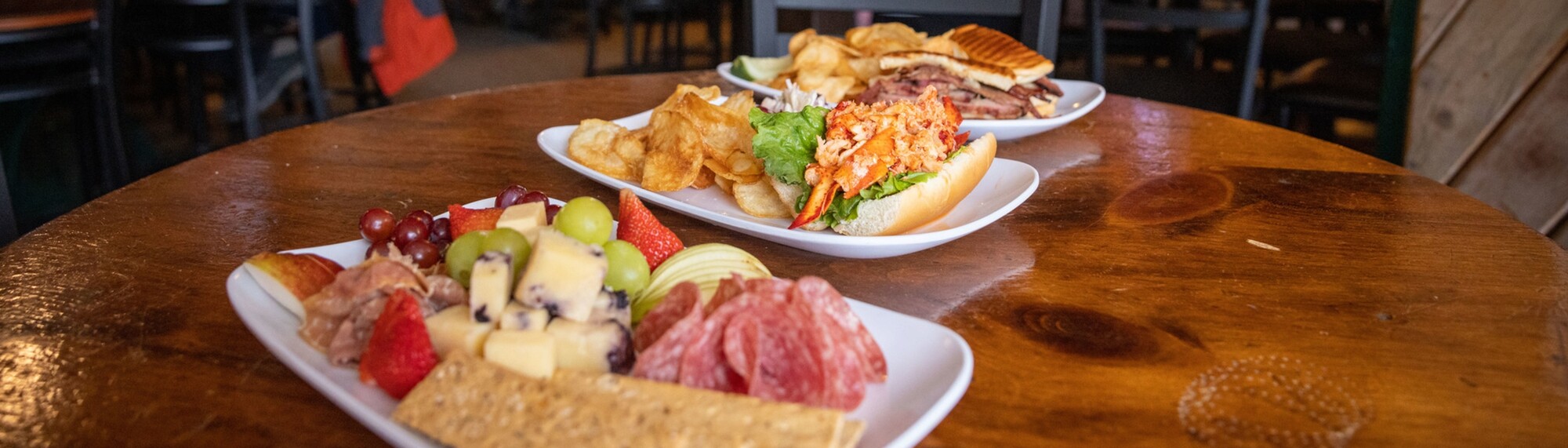 Three plates of assorted food: meats, cheeses, fruits, chips, and a lobster roll on a wooden table.