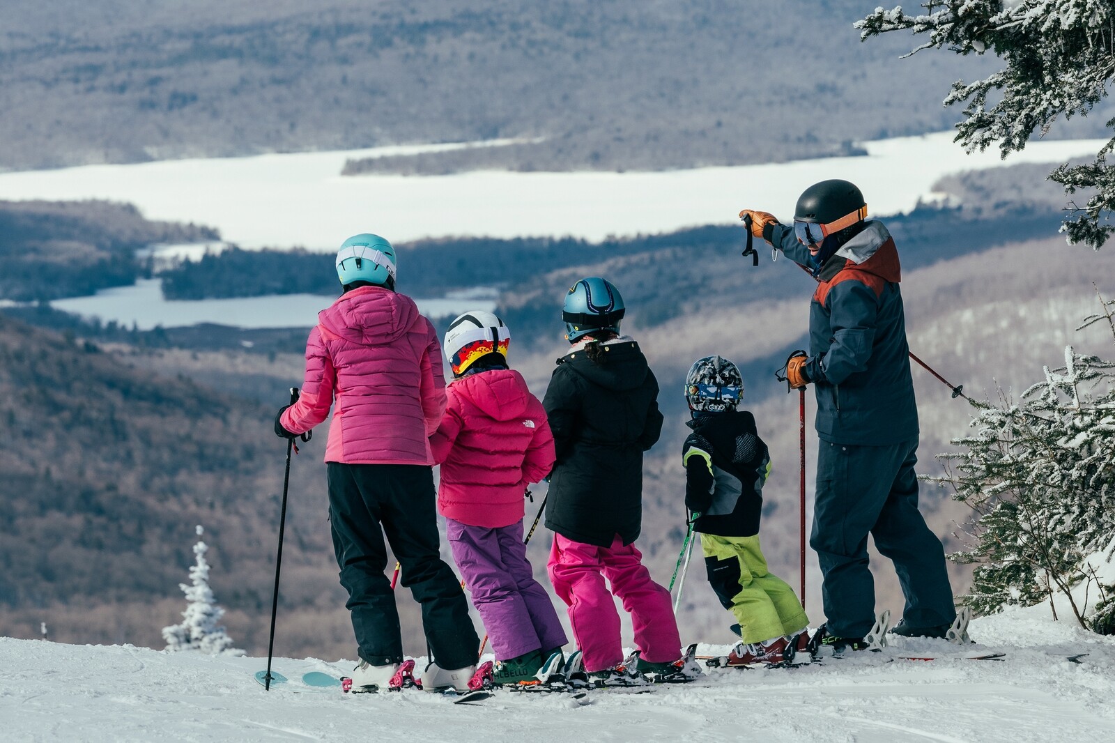 Five skiers, including children, looking at a snowy landscape from a mountain slope.