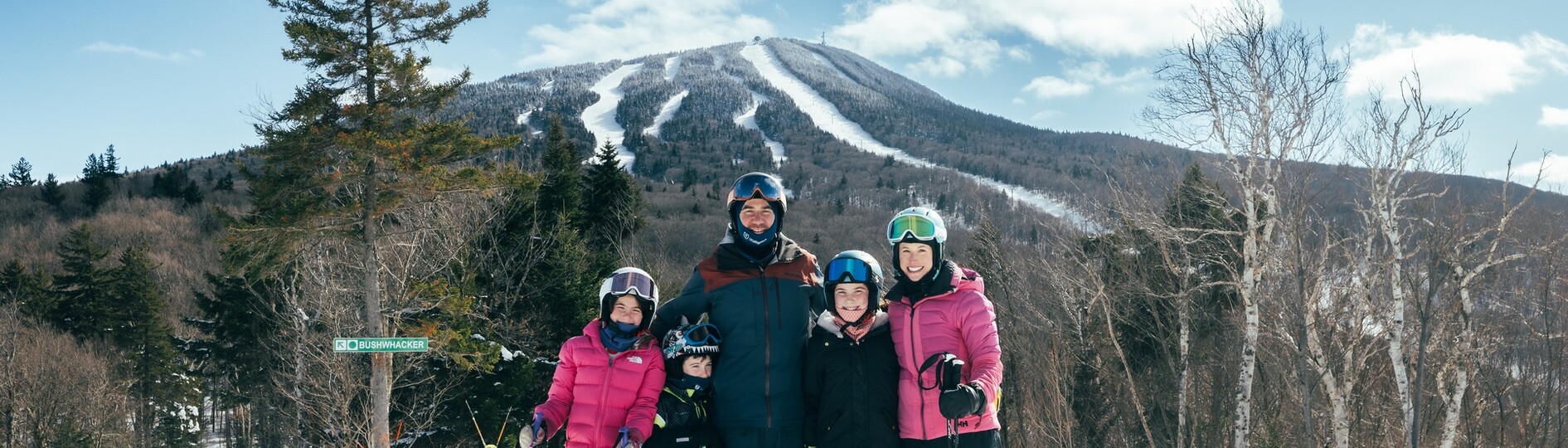 A family in ski gear poses with a mountain and ski slopes in the background.