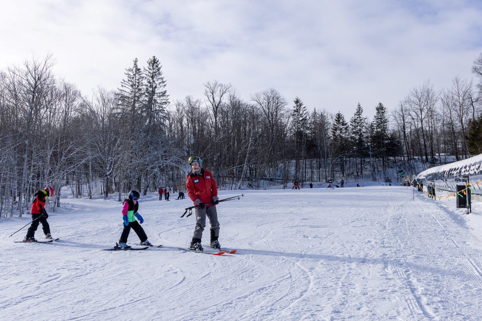 Children ski on a snowy slope, guided by an instructor in a red jacket.