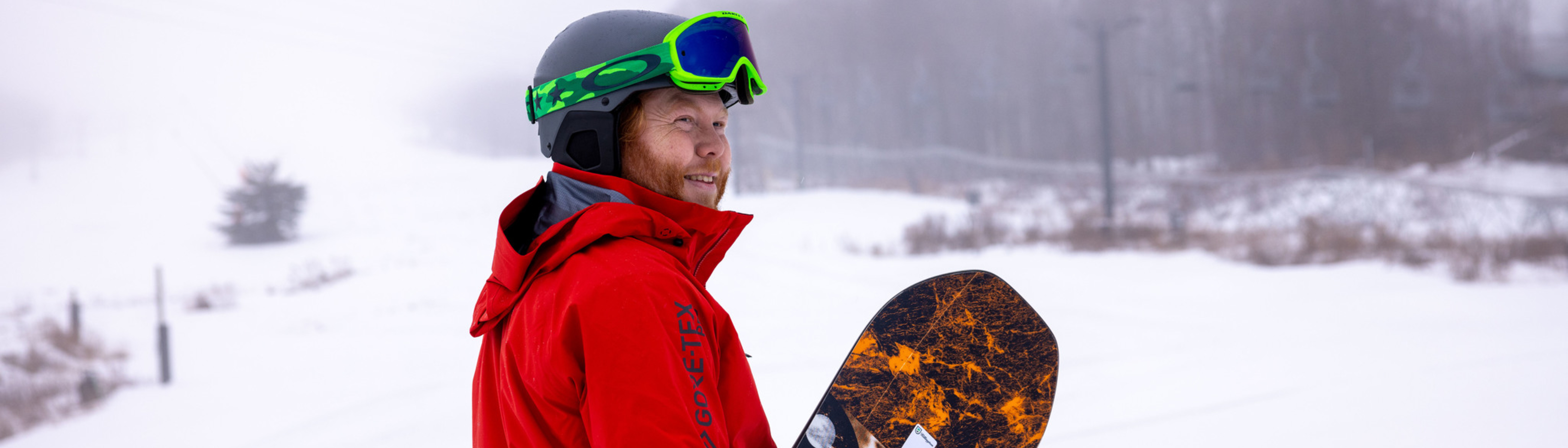 A smiling snowboarder in a red jacket and helmet stands in a snowy landscape.