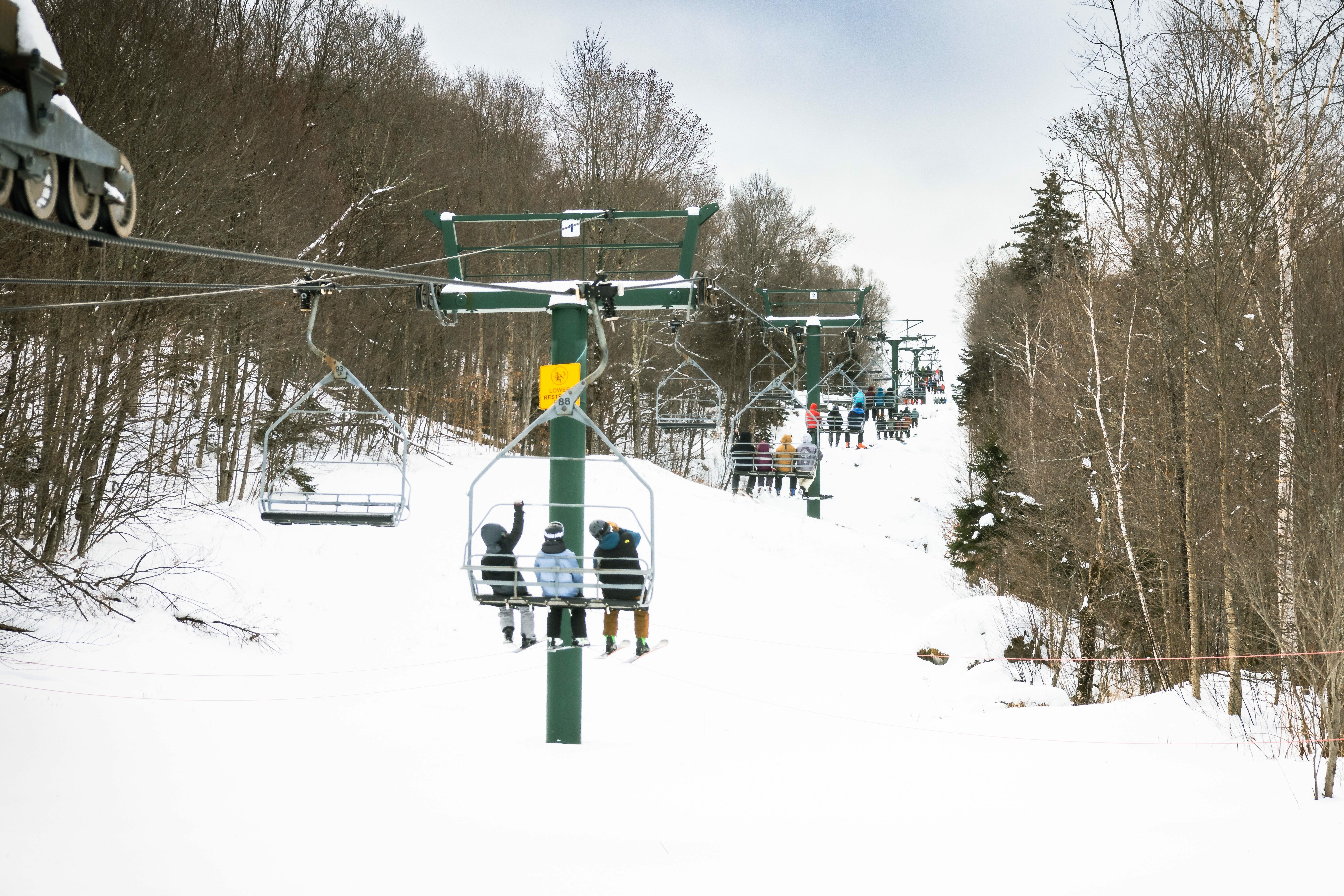 Ski lift with riders in a snowy landscape, people waiting in line below.