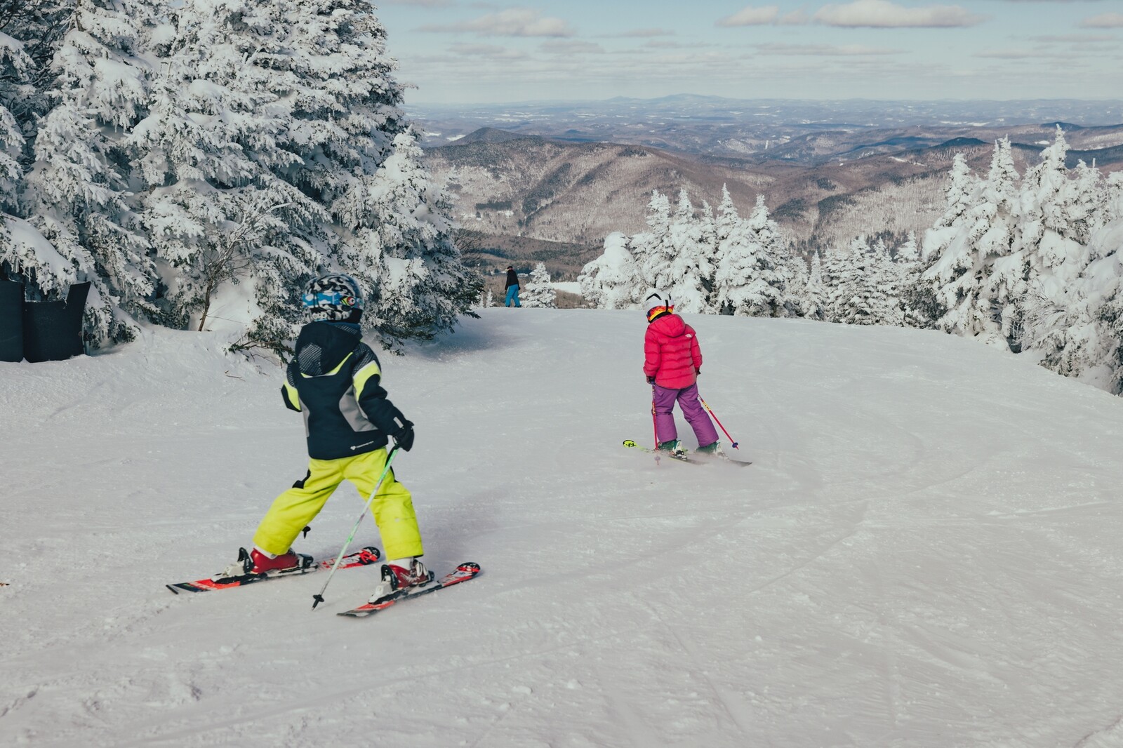 Two children skiing down a snowy slope surrounded by trees and mountains.
