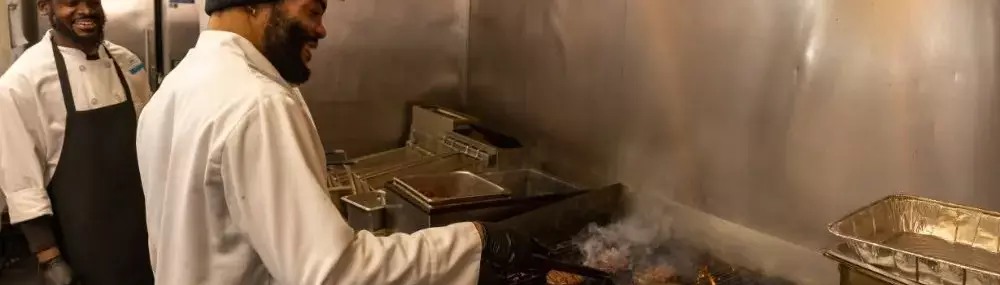 Two chefs cooking on a grill in a kitchen, with steam rising from the food.