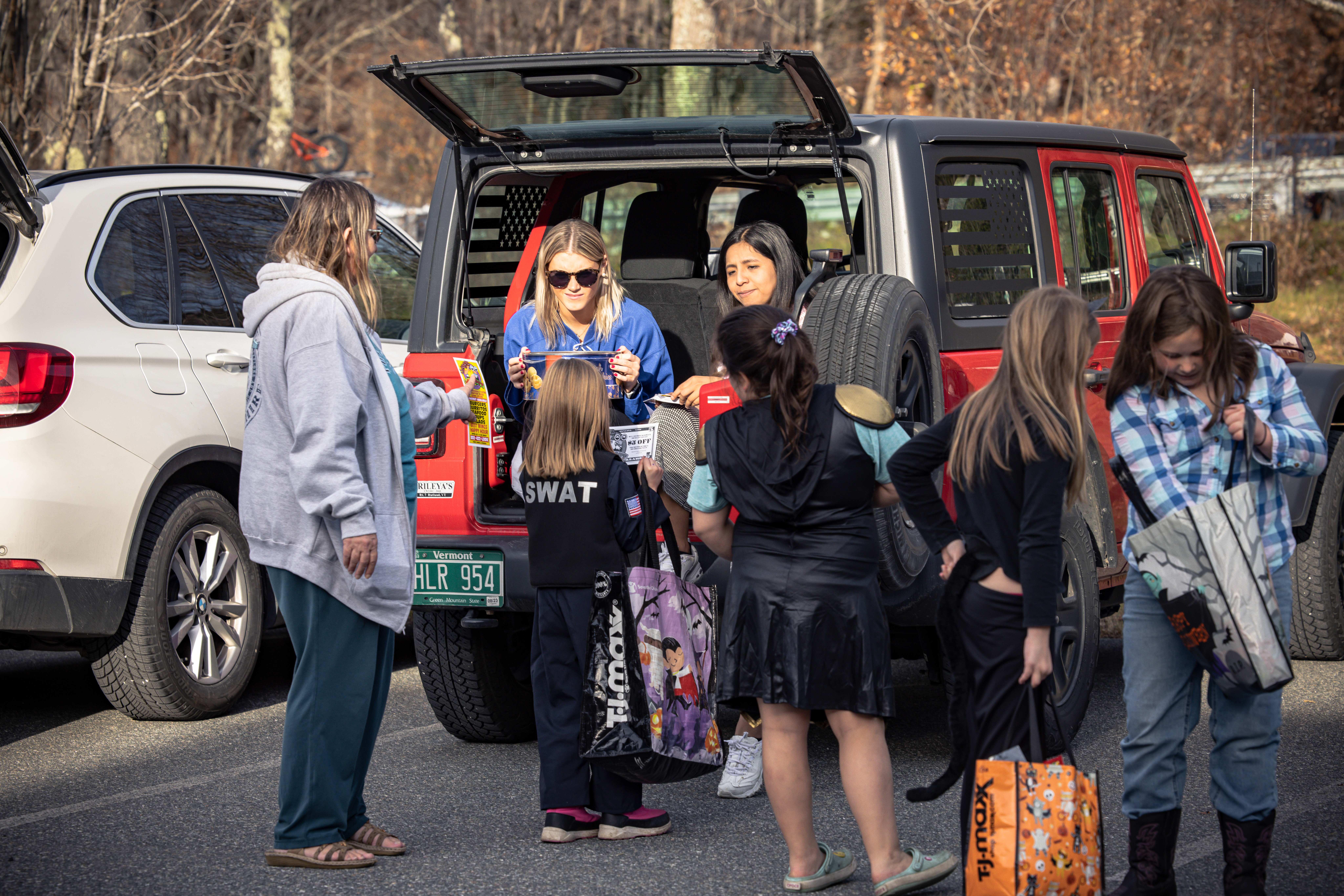 Children in costumes gather around a car, receiving treats from two smiling adults.