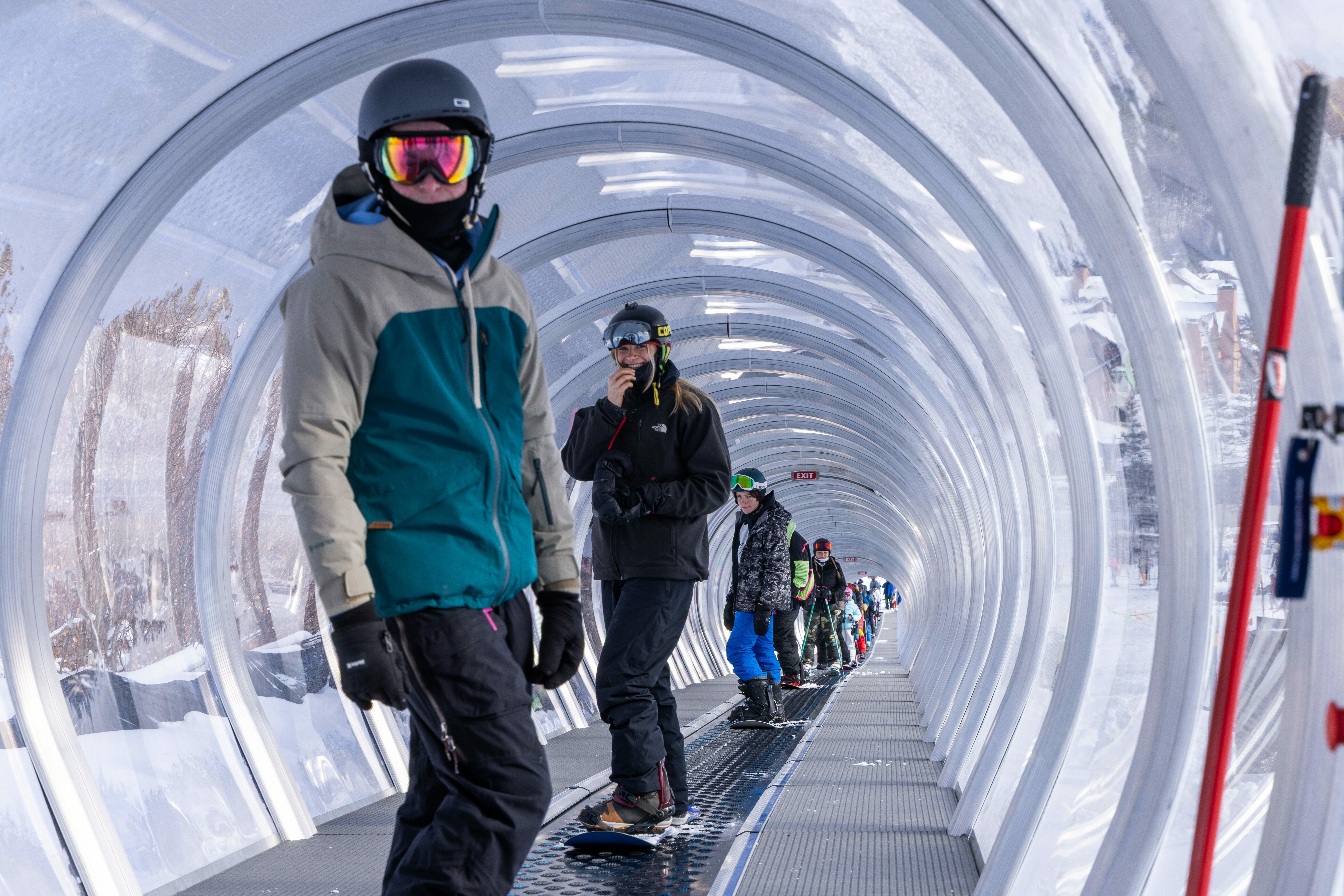 Snowboarders waiting in a clear, enclosed lift tunnel at a ski resort.