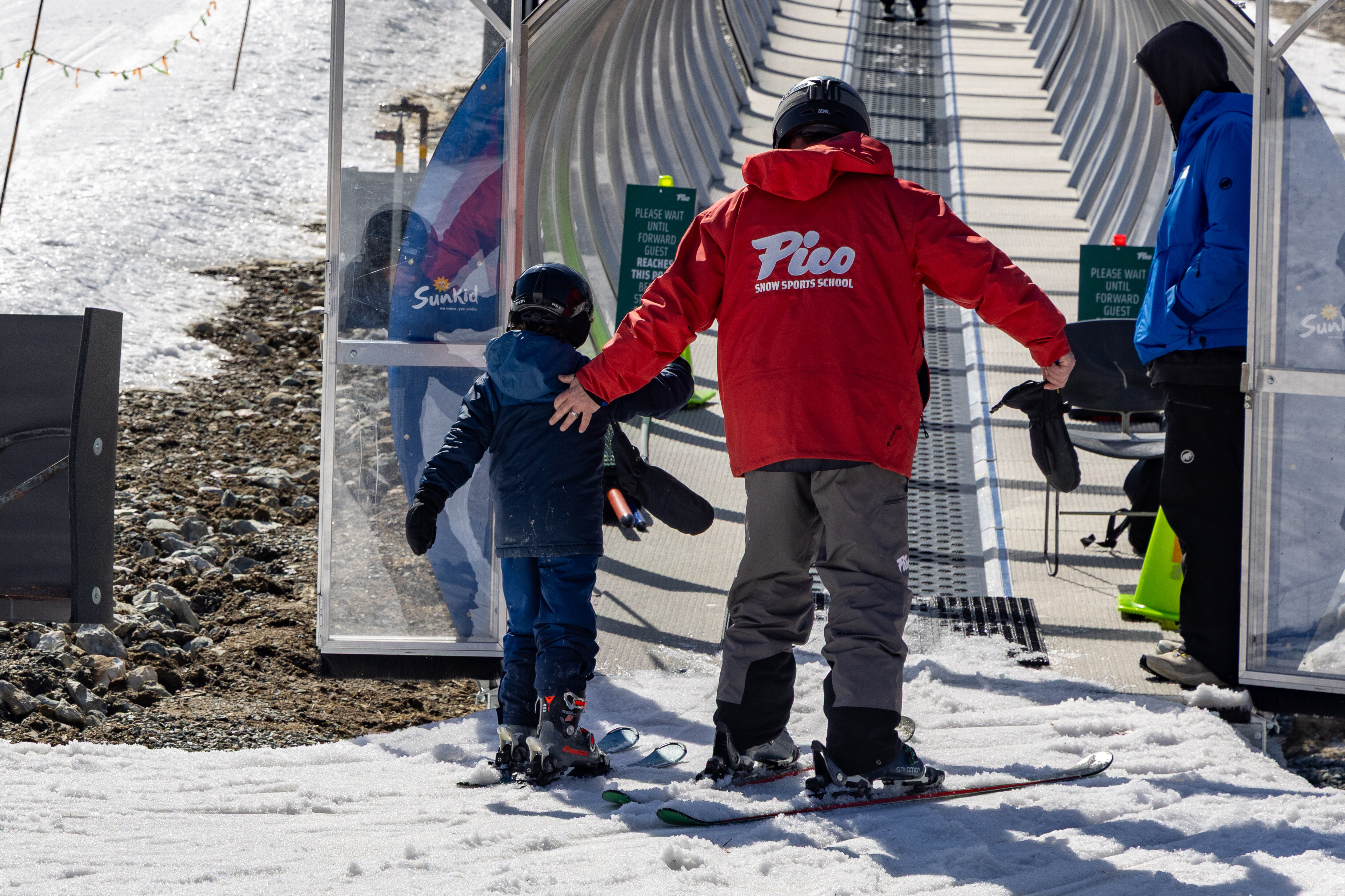 A ski instructor guides a child towards a ski lift on snow-covered terrain.