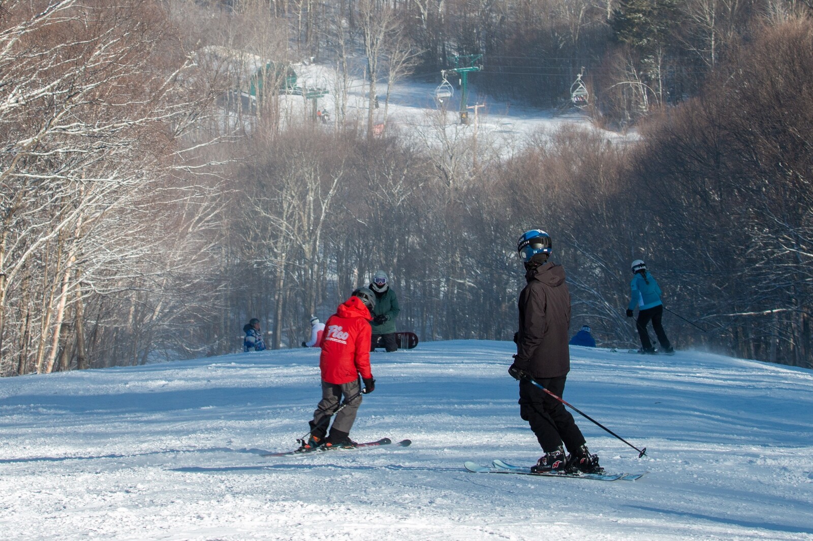 Two skiers interact on a snowy slope, with others skiing in the background.