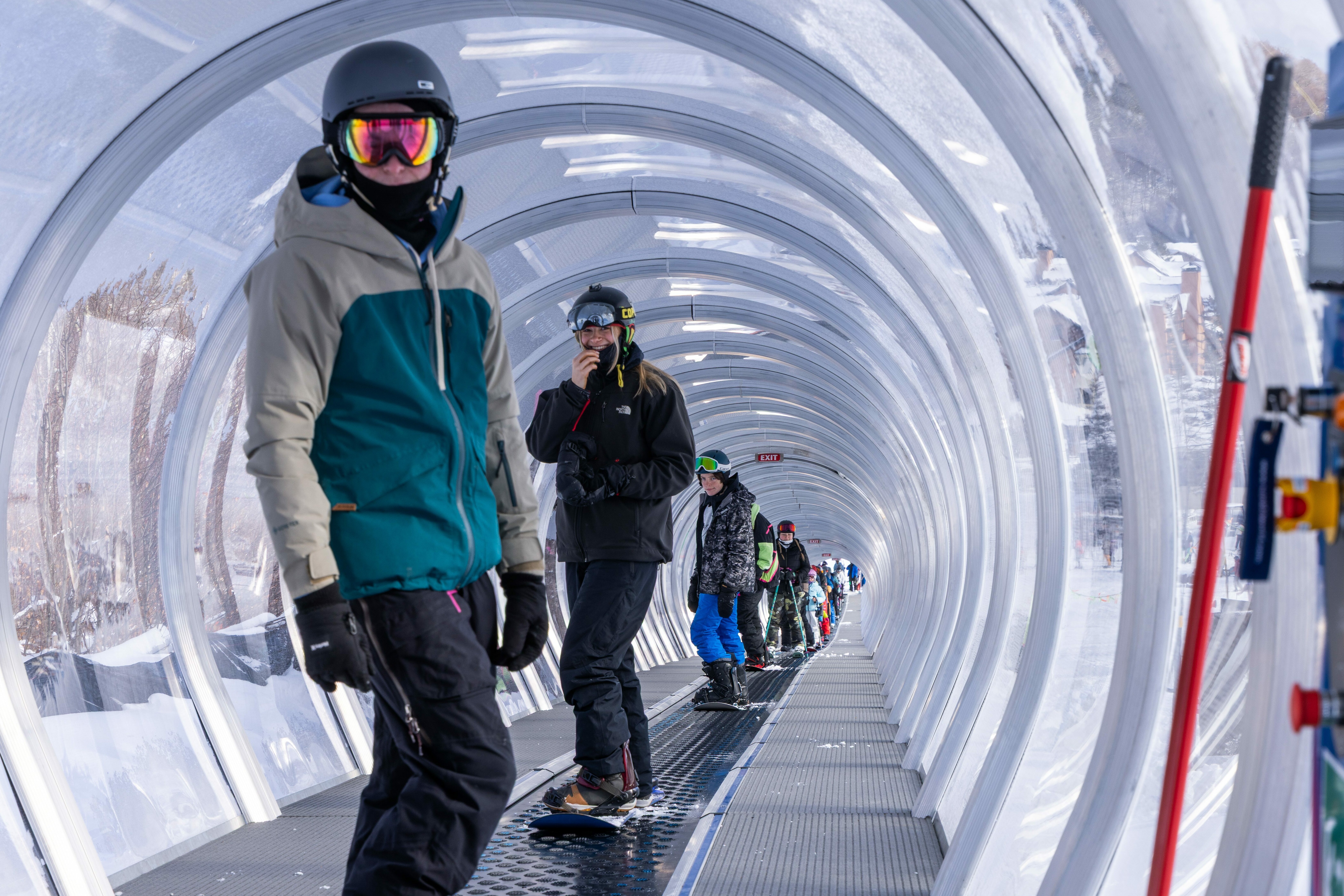 Snowboarders in a transparent tunnel moving up a ski slope.