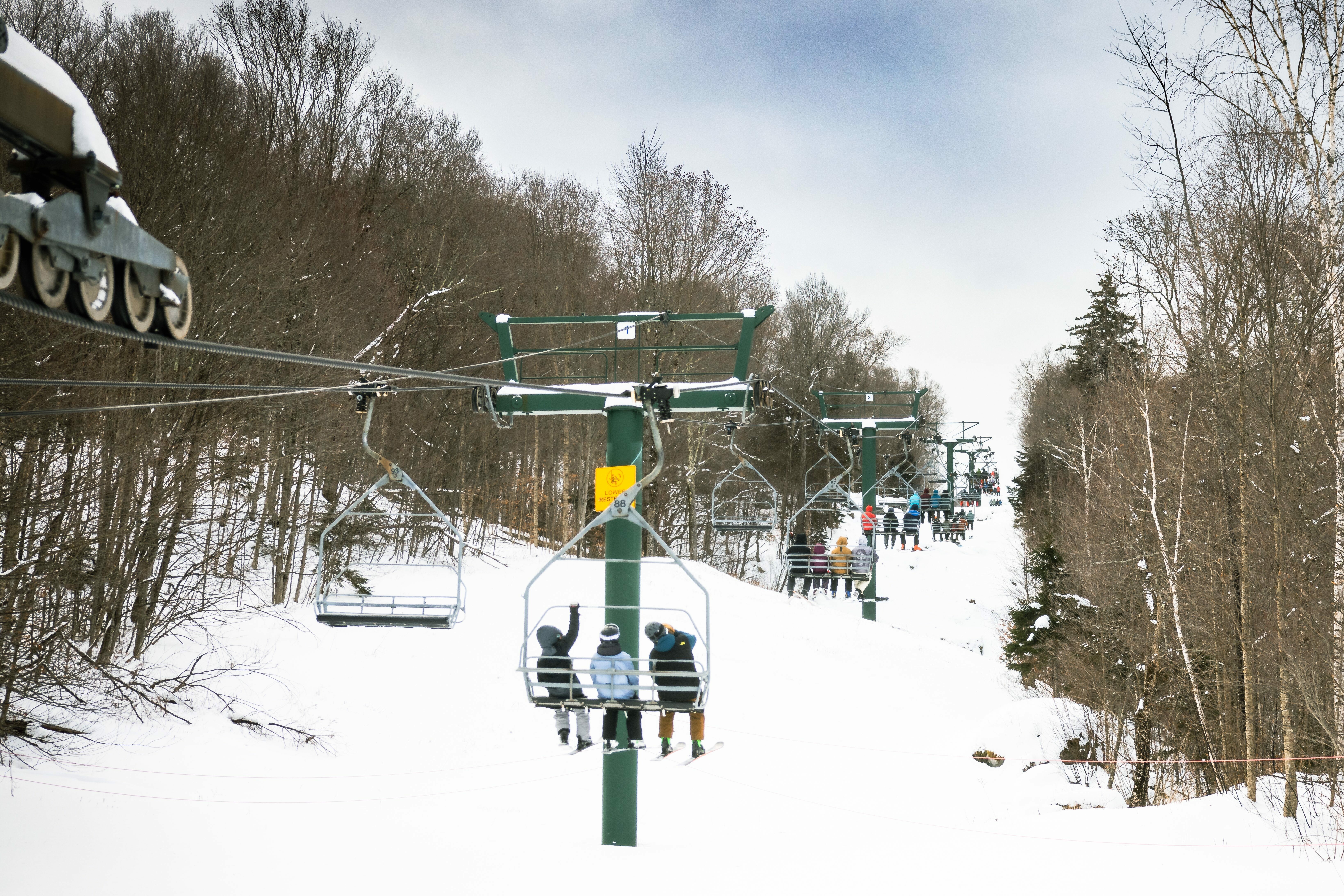 Skiers on a lift with snowy slopes and trees in the background.