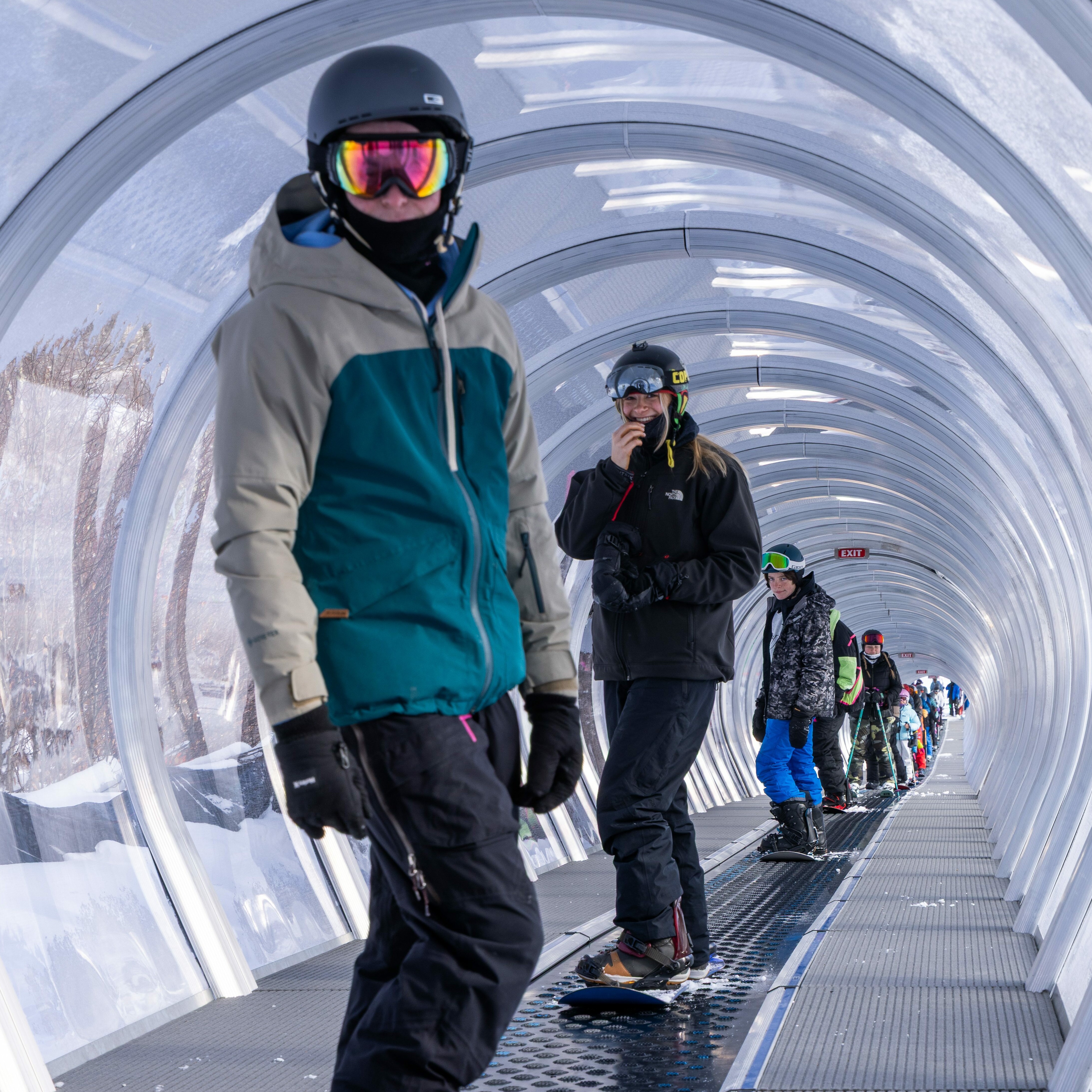 Snowboarders in helmets and gear walking through a covered snow tunnel.